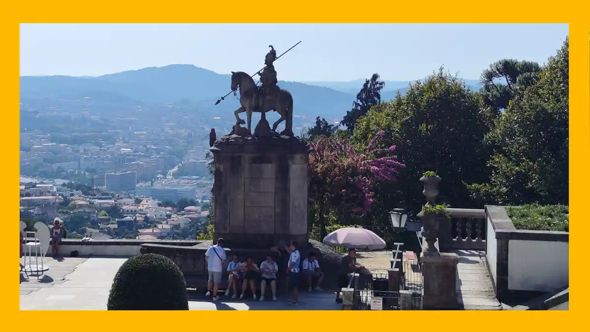 Estátua equestre em granito de São Longuinho no Santuário do Bom Jesus do Monte, com visitantes no terreiro e a vista panorâmica da cidade de Braga ao fundo.
