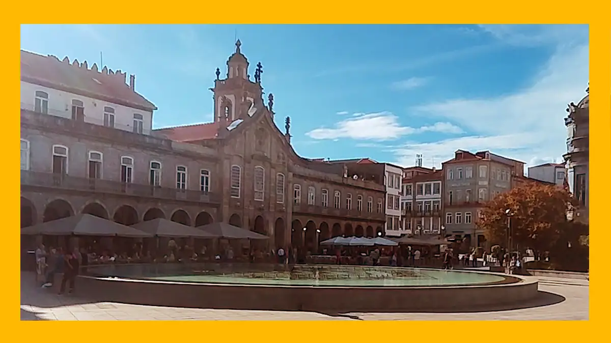 Imagem com moldura amarela mostrando o chafariz da Praça da República em Braga com água, as esplanadas da Arcada e a Igreja da Lapa sob um céu azul ensolarado.